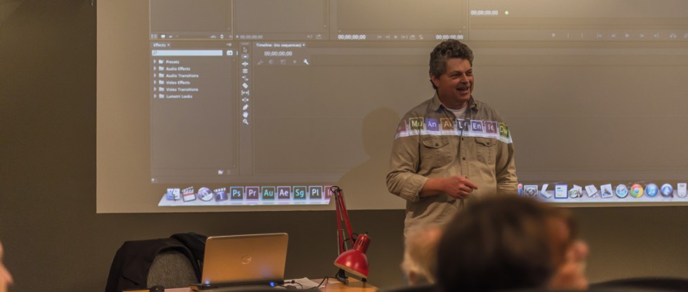 A photo of a presenter in front of a screen showing the interface of Adobe Premiere