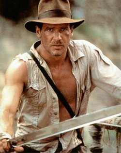 A promo photo of Harrison Ford as Indiana Jones, from Indiana Jones and the Temple of Doom. One sleeve of his shirt is torn, and he is holding a machete towards the camera.