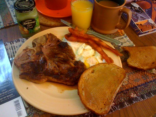 A pale yellow plate filled with a t-bone steak, bacon, eggs over easy, and toast.
