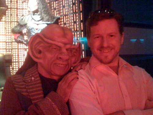 A photo of me from Star Trek: The Experience in Las Vegas. I am wearing a white shirt and have sunglasses propped on my head. A roving actor dressed as a Ferengi alien, with a large wrinkled head and enormous ears, rests his hand on my shoulder. Behind us is a museum case with some Klingon costume props from Star Trek.