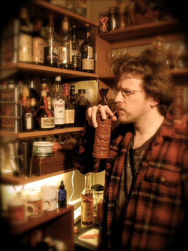 A picture of me in front of the tiny hanging shelf that used to be my "tiki nook" bar area, with tiki mugs on the shelves. I am wearing a red flannel shirt and sipping on a straw in a tiki mug