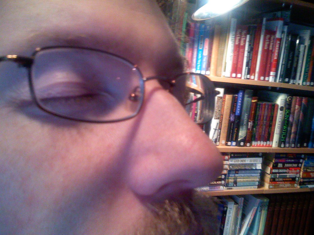 An extreme close-up of a bespectacled man's face in profile, eyes closed, in front of shelves packed with books and DVDs.